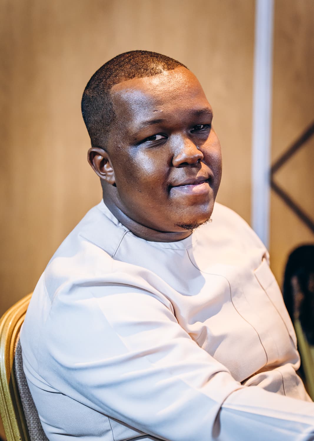 A smiling Black man with short hair wearing a light beige long-sleeved shirt indoors.
