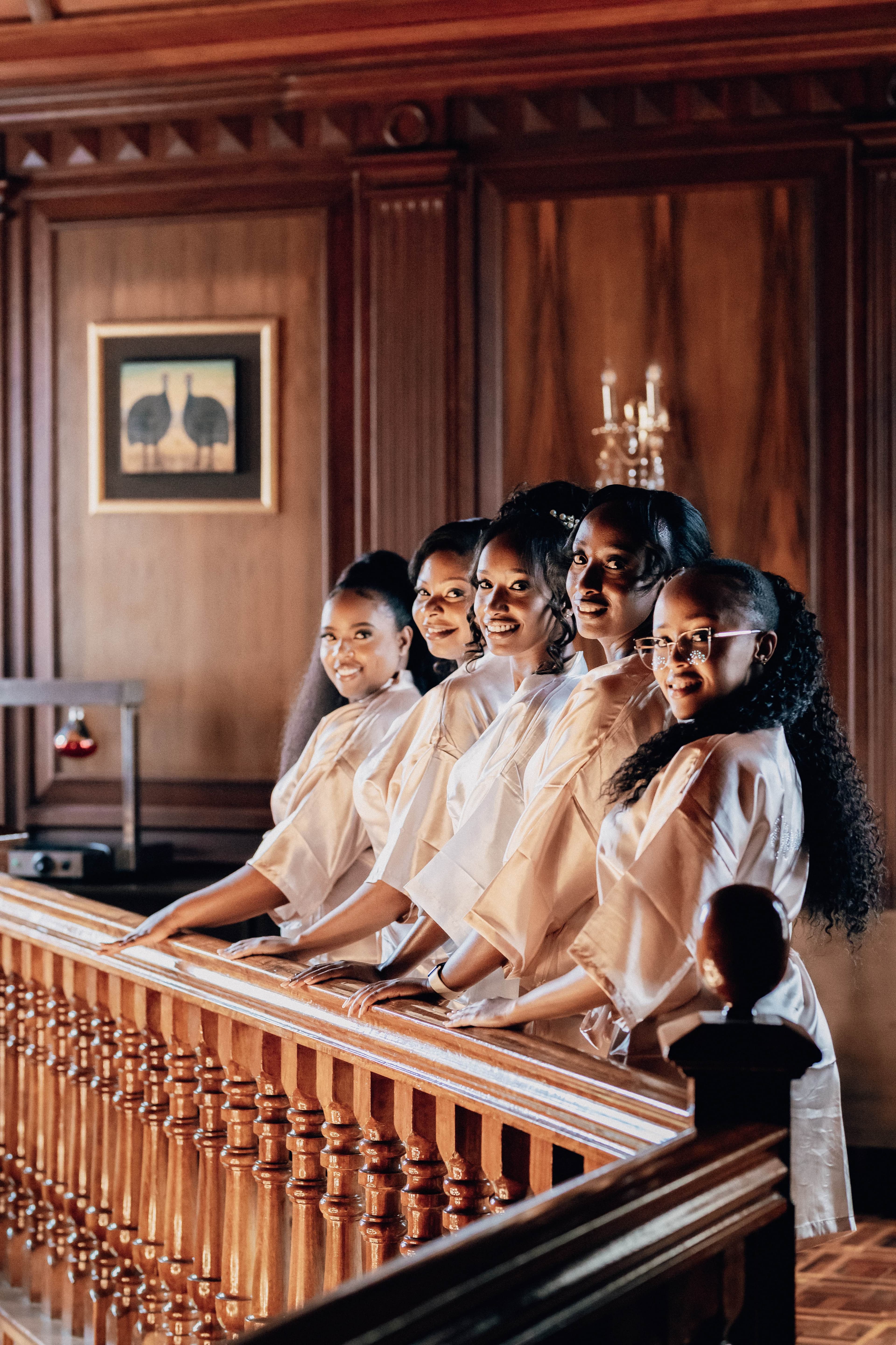 Five women in white satin robes smile while leaning against a wooden staircase railing.