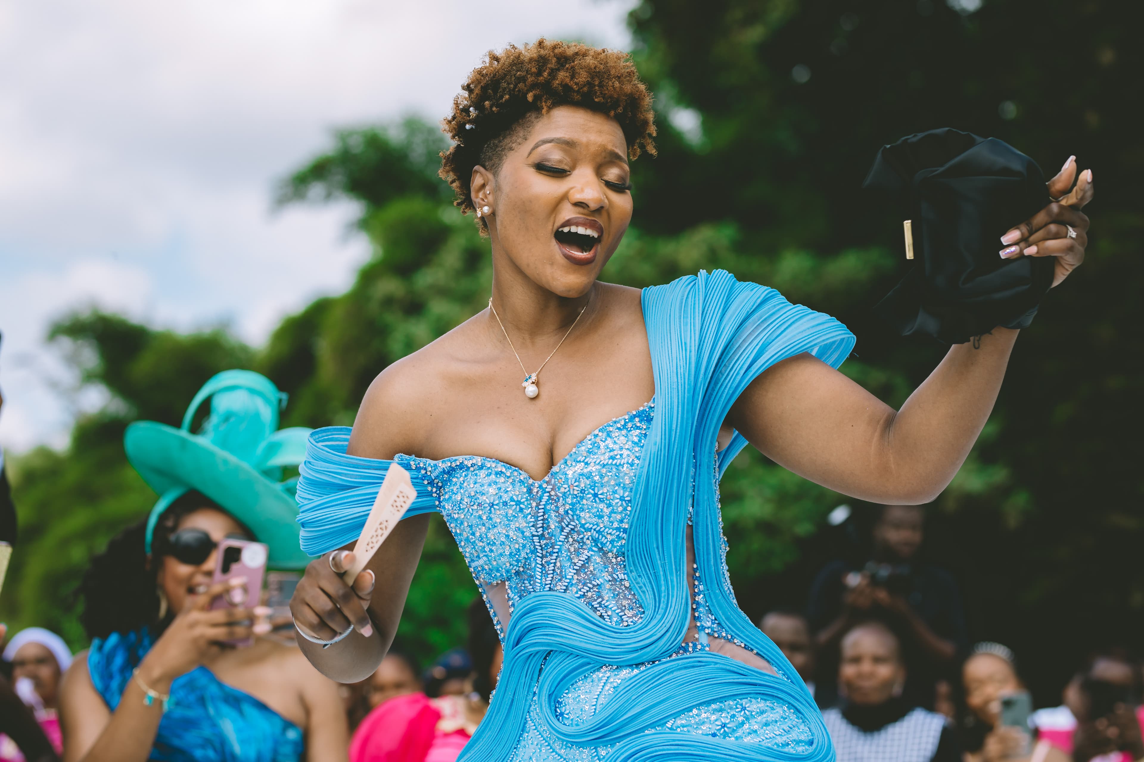 Joyful woman in a vibrant blue sequined gown dances holding a fan and black clutch.