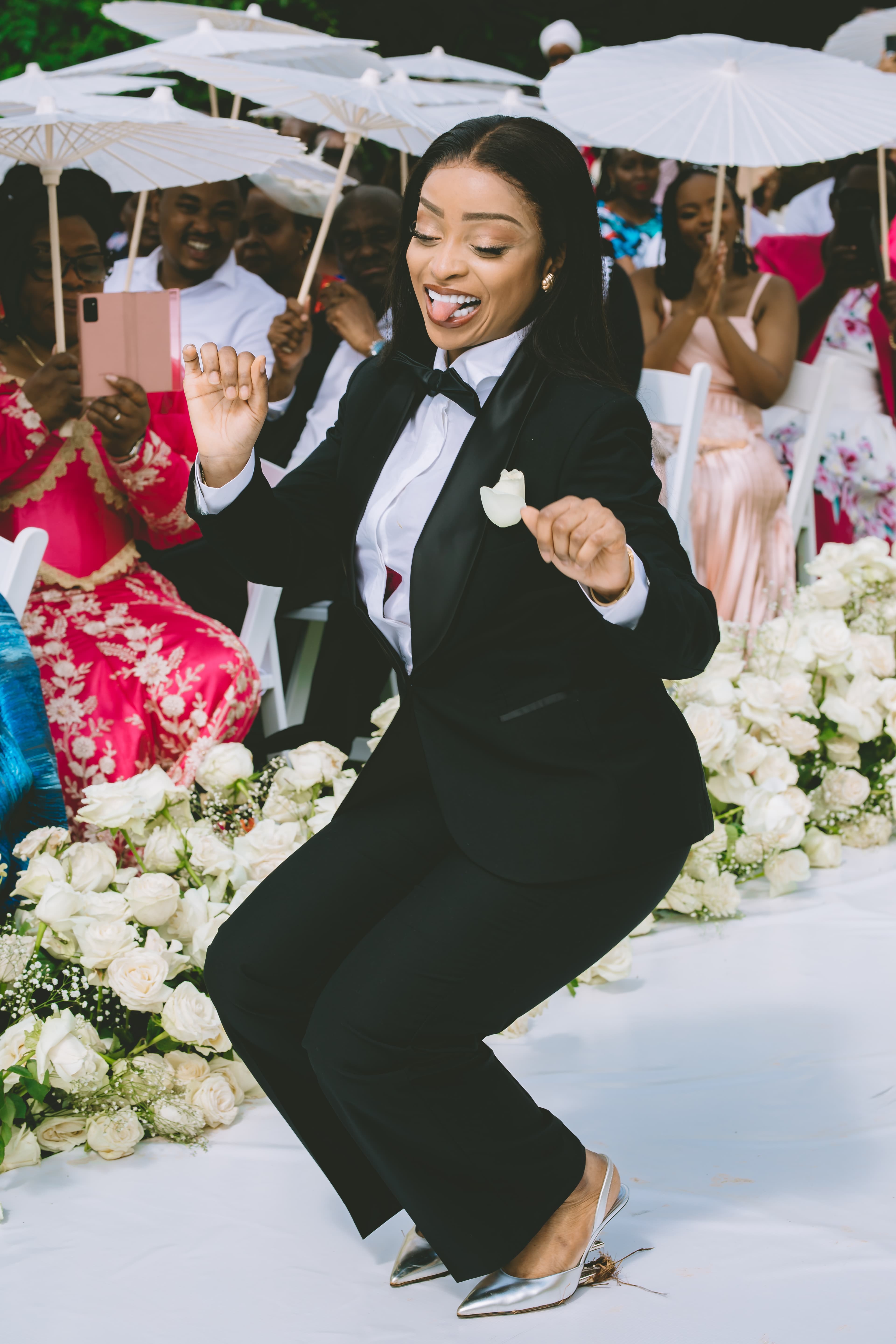 Woman in a black tuxedo dancing playfully at an outdoor wedding with white roses.
