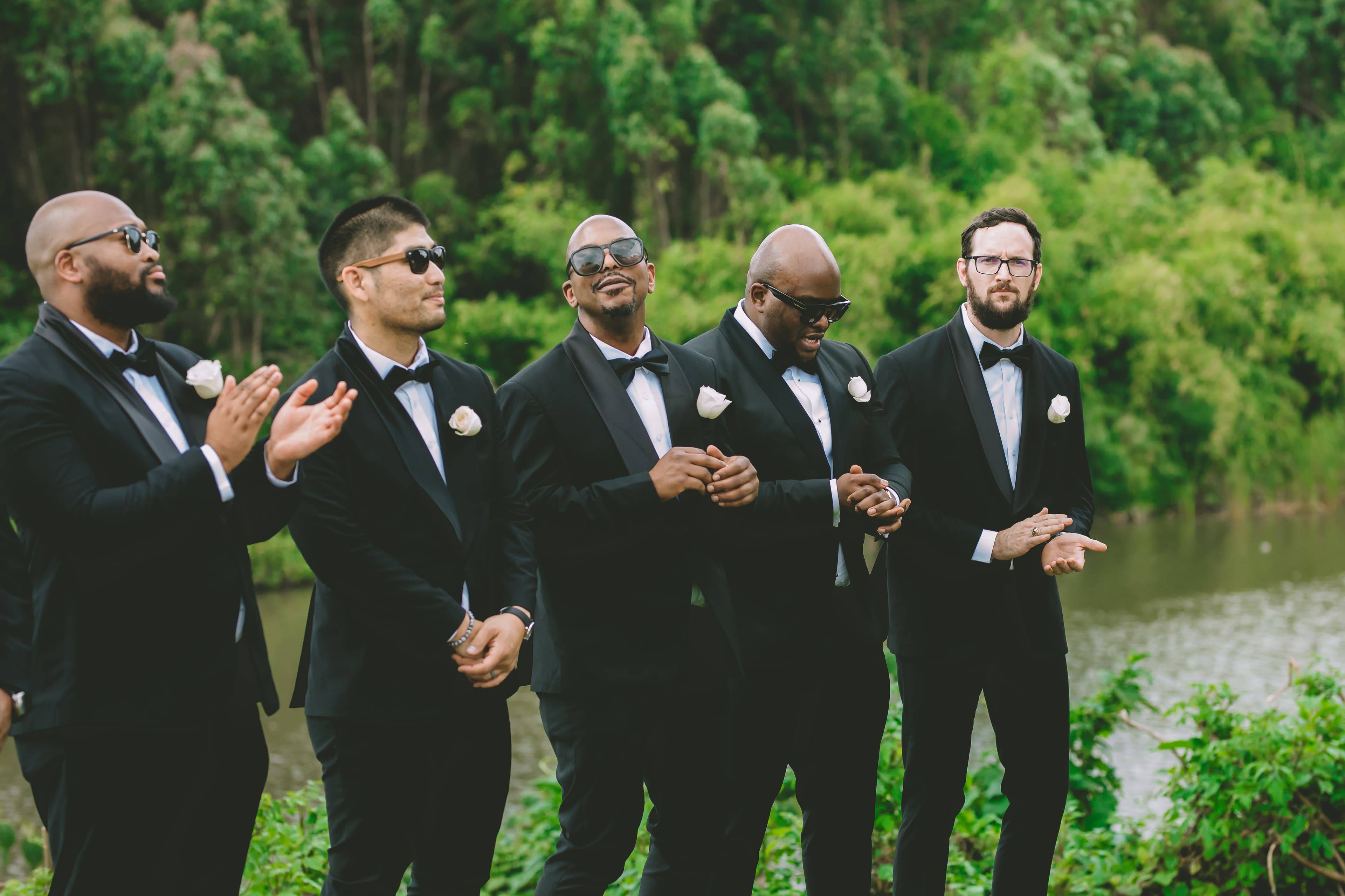 Five groomsmen in black tuxedos stand outdoors near water, some wearing sunglasses and clapping.