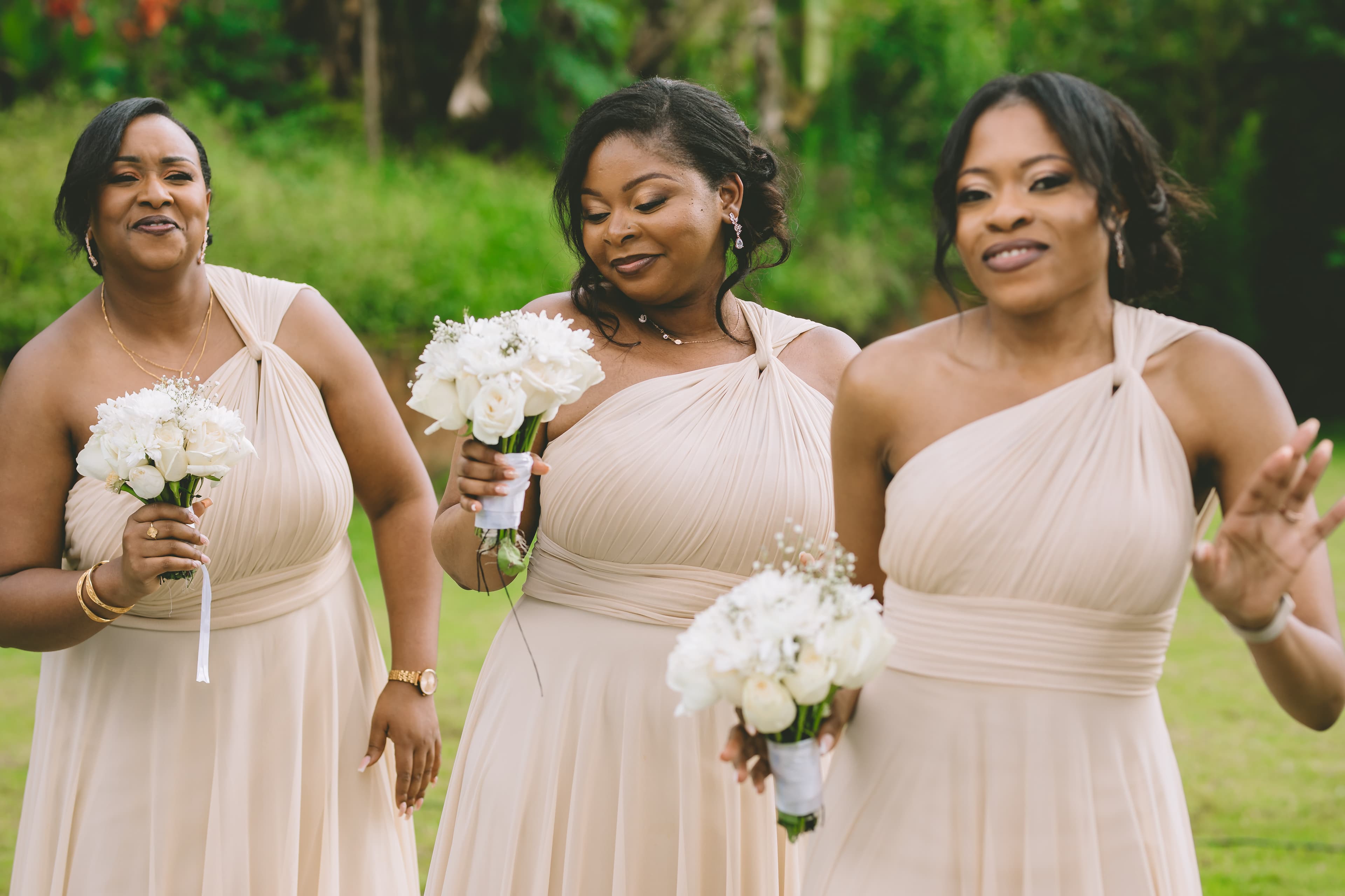 Three bridesmaids in beige one-shoulder dresses holding white rose bouquets in a green garden.