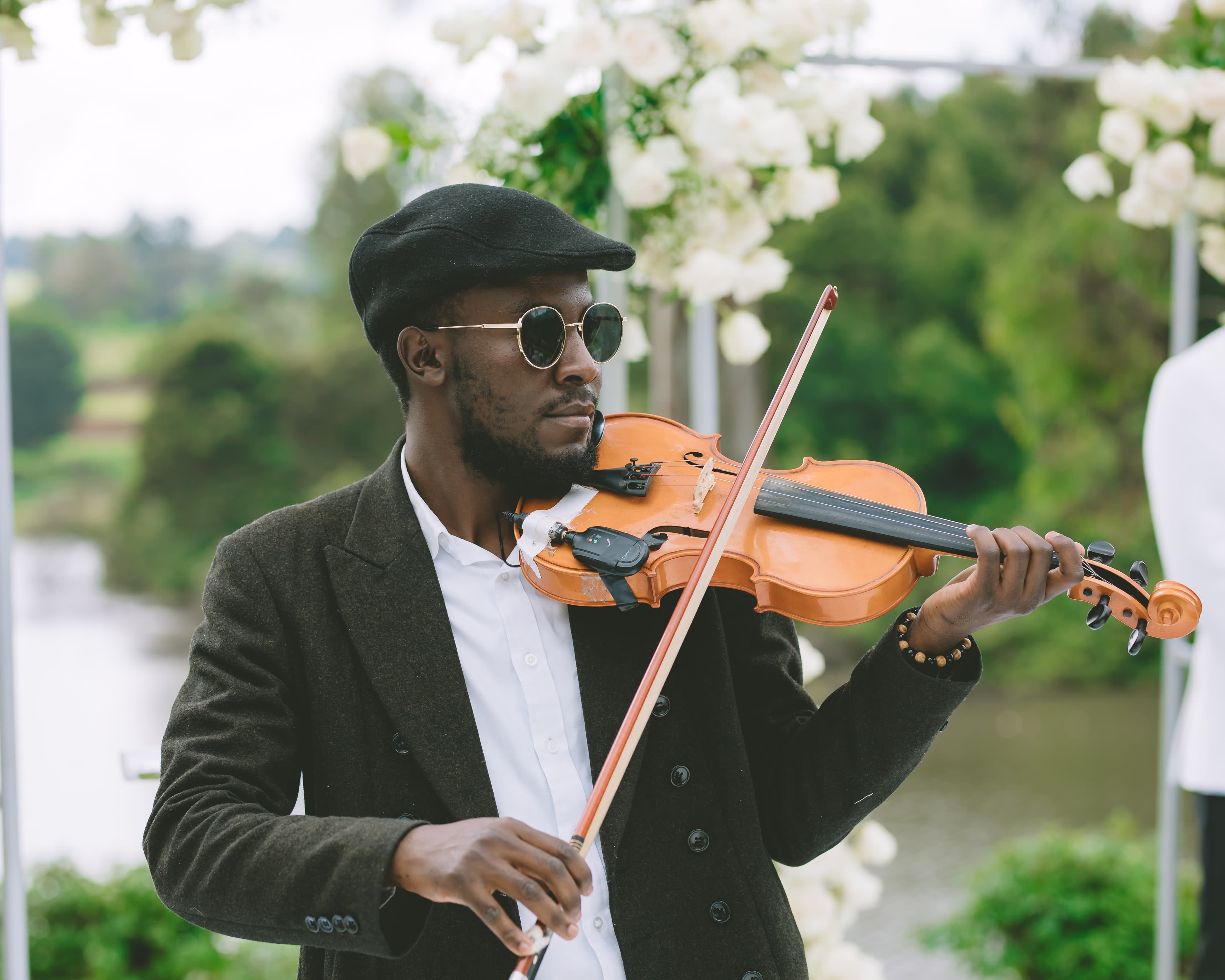 Black man in a flat cap and sunglasses playing violin outdoors near white wedding flowers.