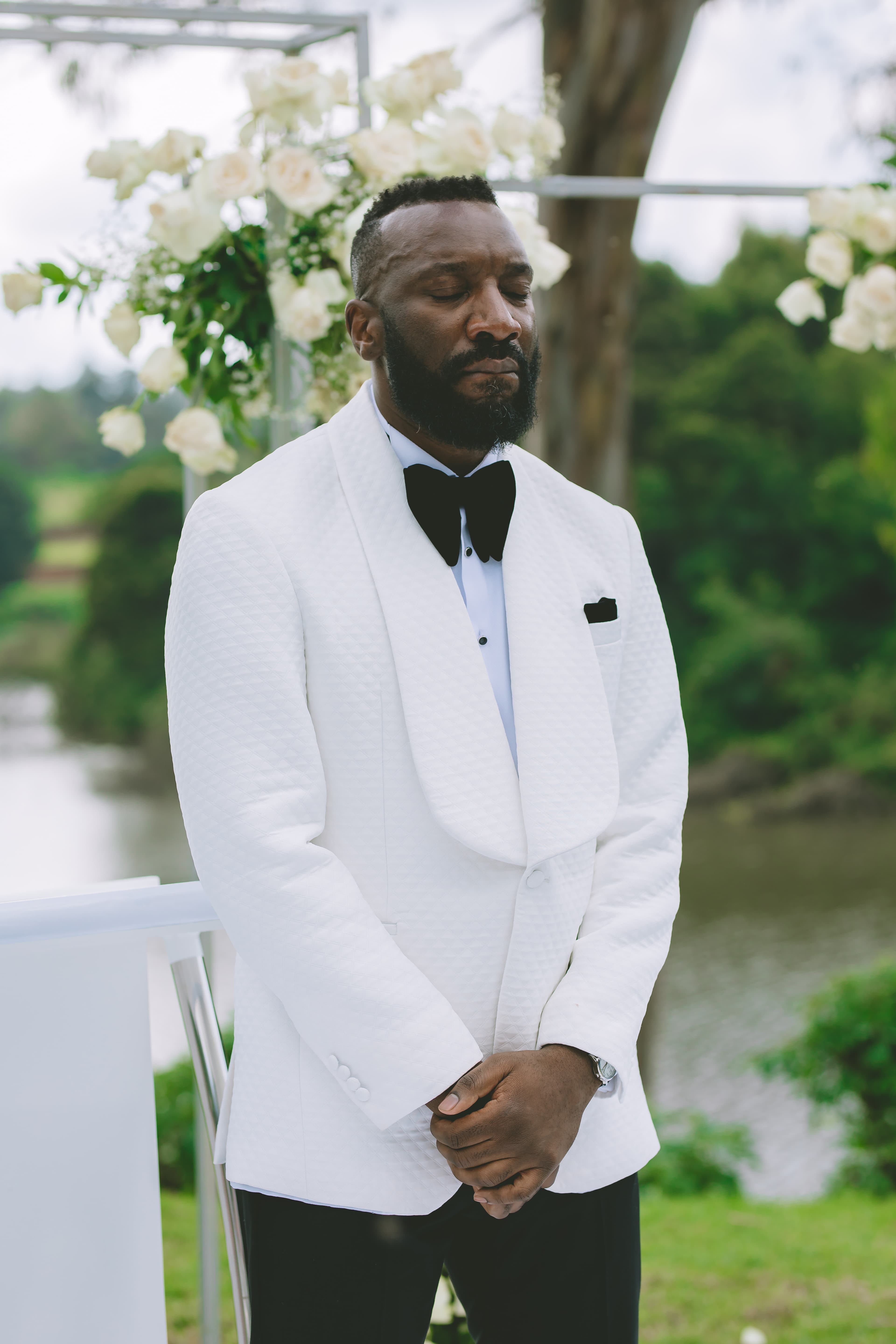 Bearded Black man in white textured tuxedo jacket with eyes closed at outdoor wedding.