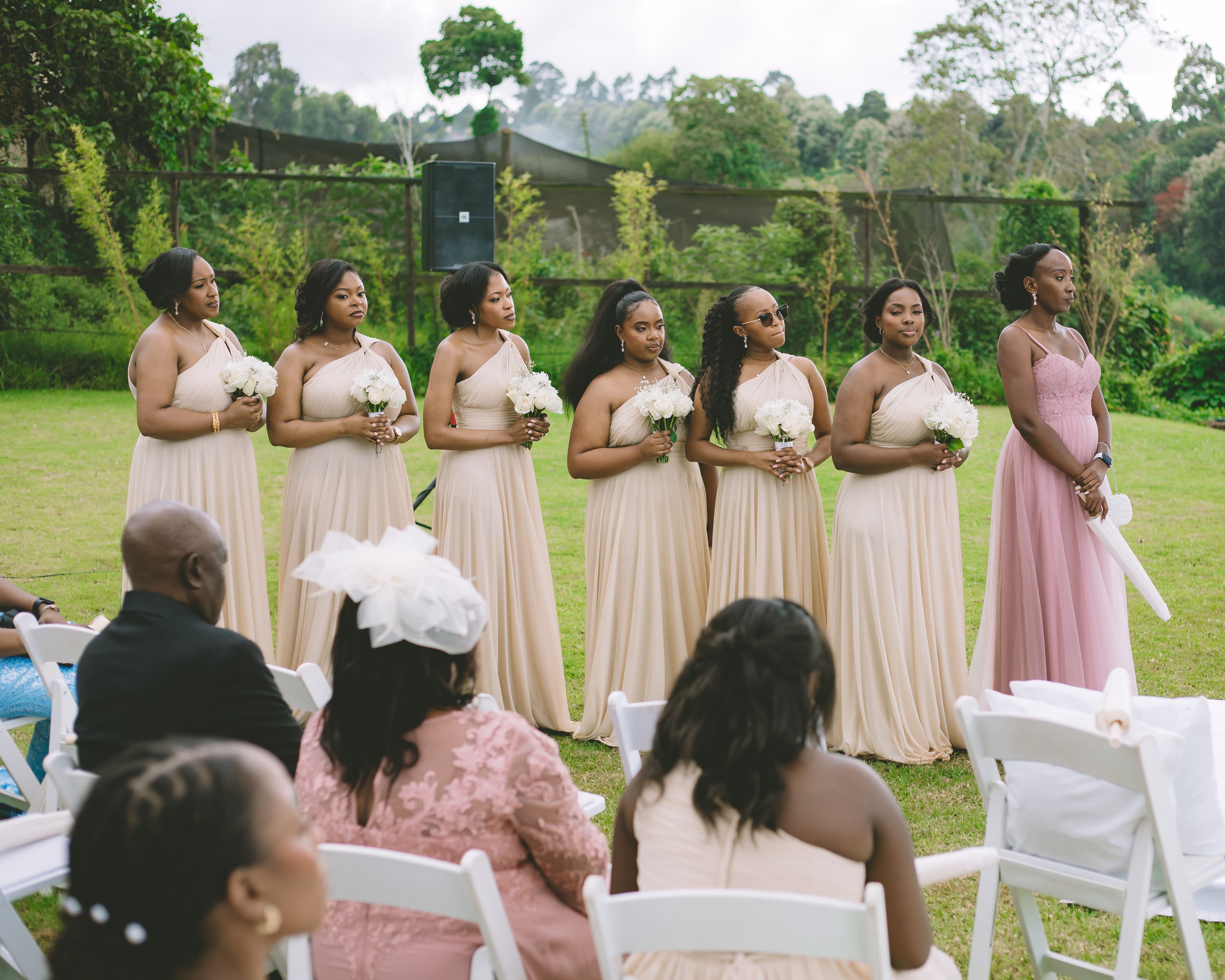 Six bridesmaids in beige dresses and one in pink stand at an outdoor wedding ceremony.
