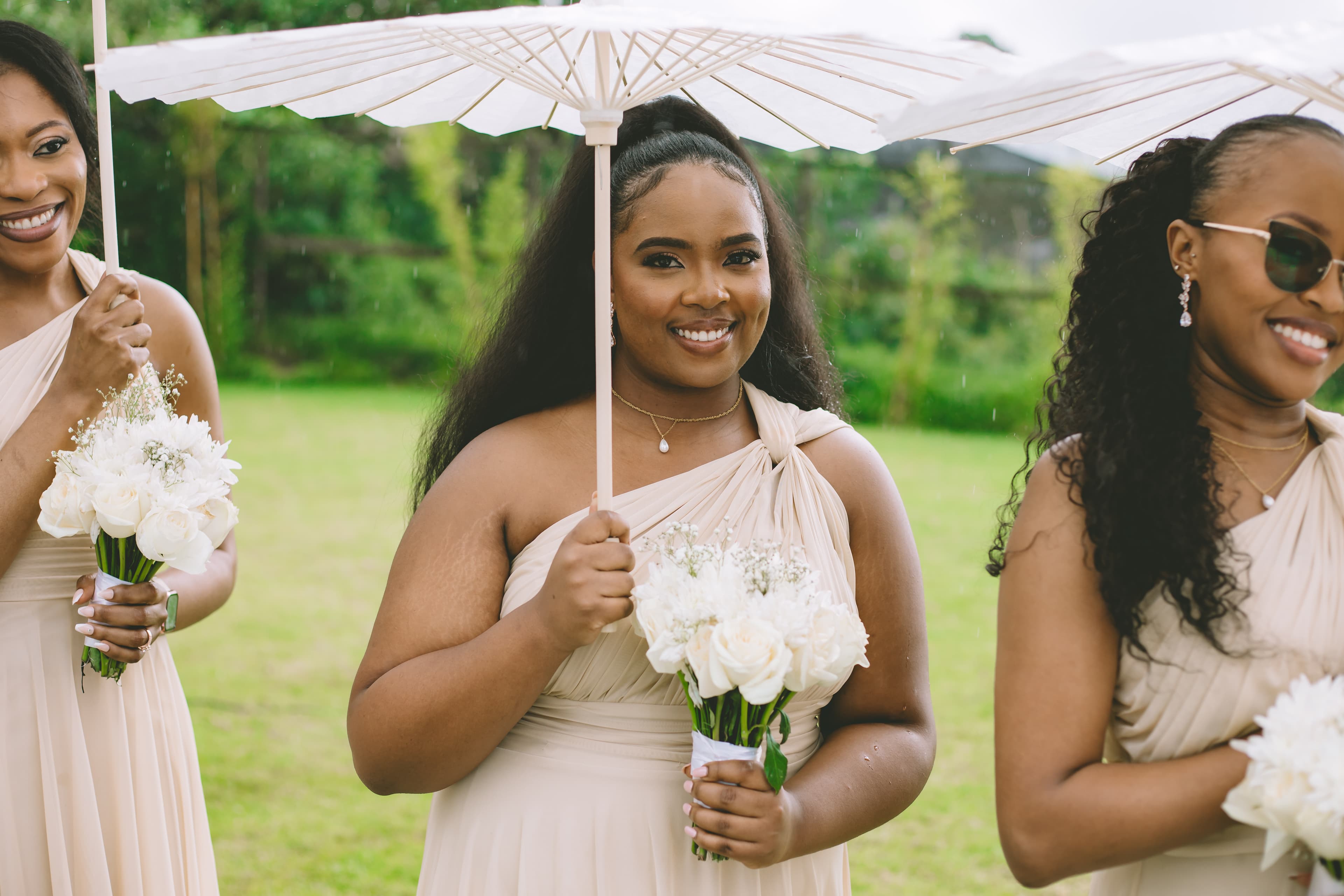 Three smiling bridesmaids in beige dresses hold white bouquets and parasols in a green field.