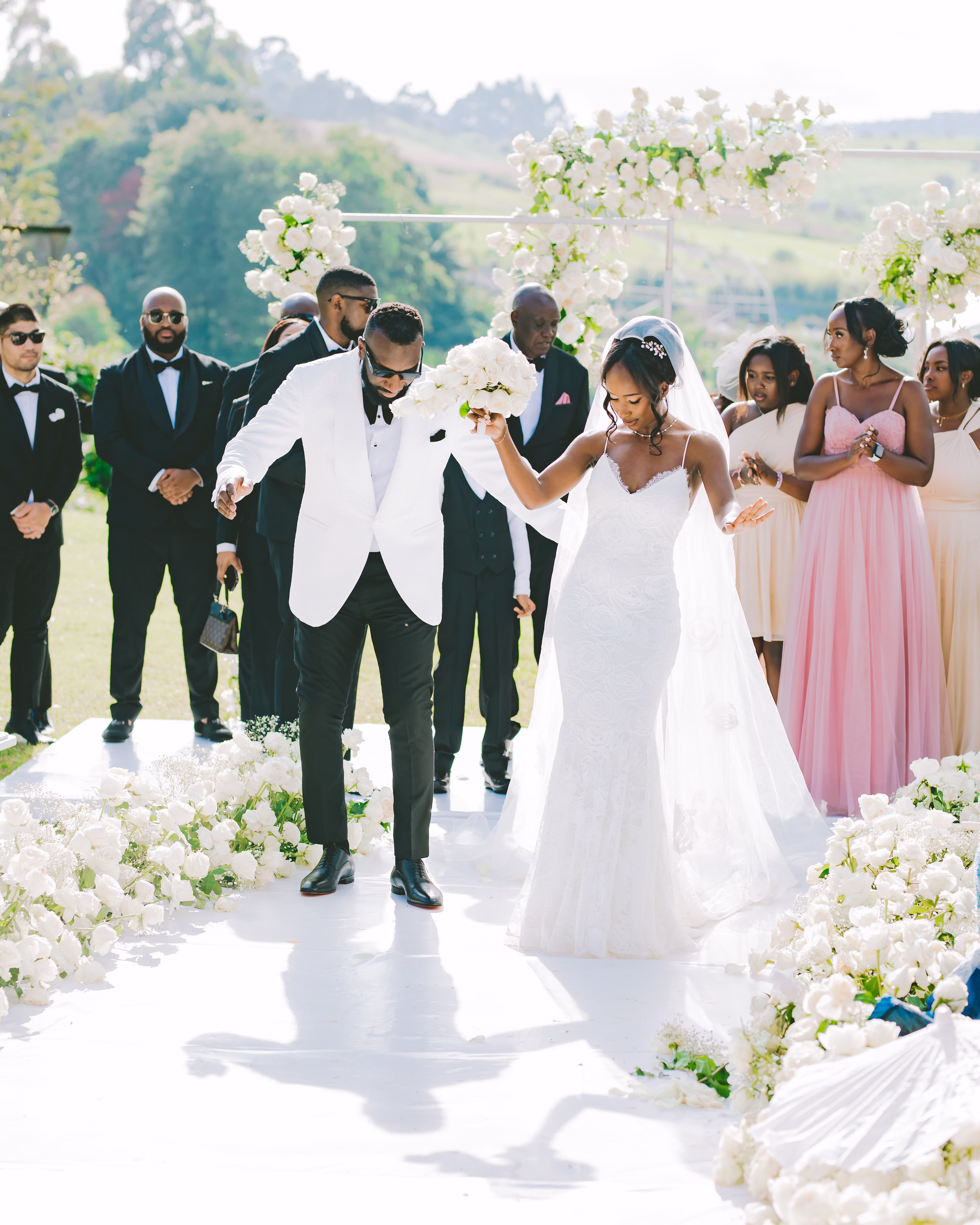 Newlyweds walk down a white floral aisle surrounded by their wedding party outdoors.