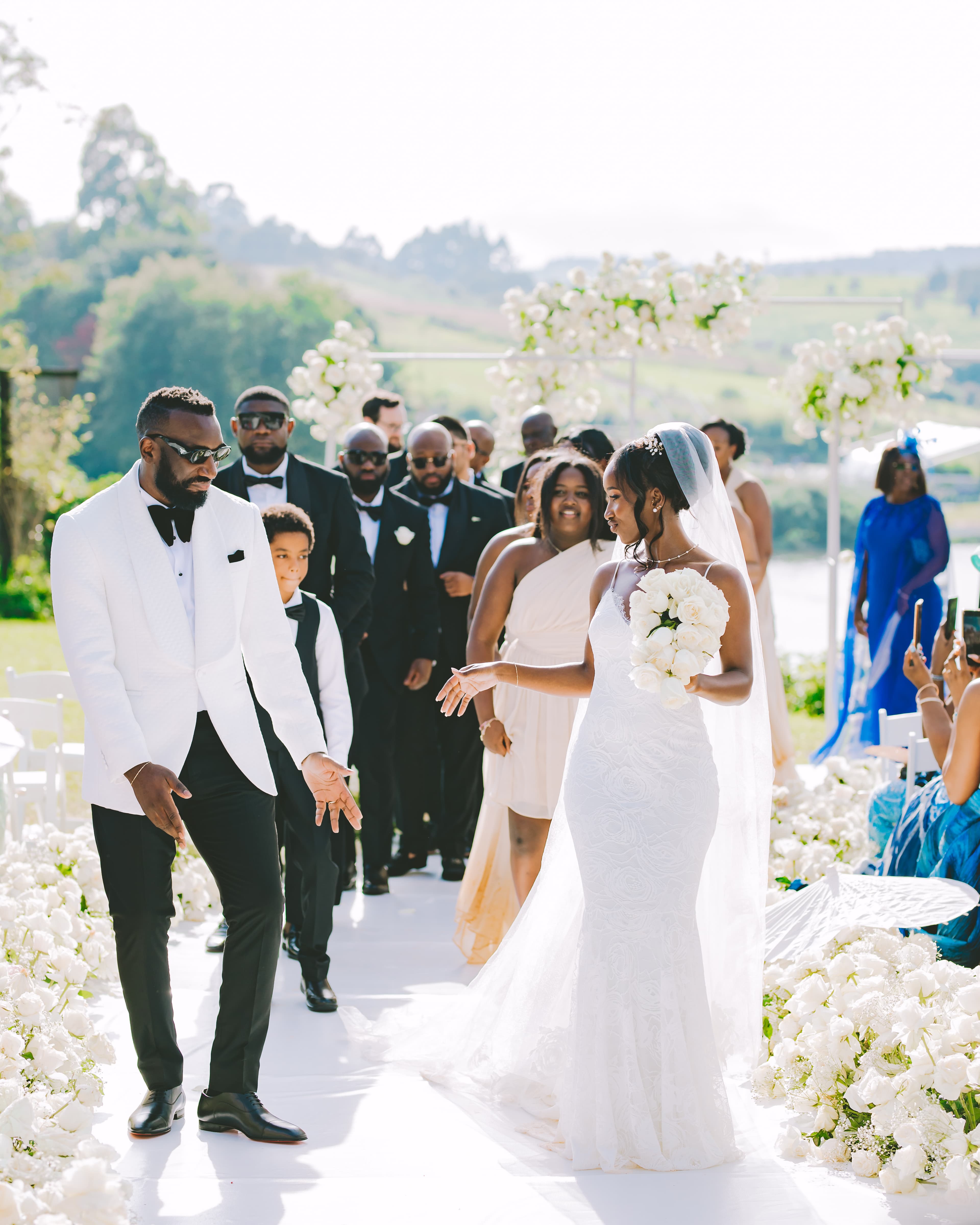 Bride and groom walk down a white floral-lined aisle during their outdoor wedding ceremony.
