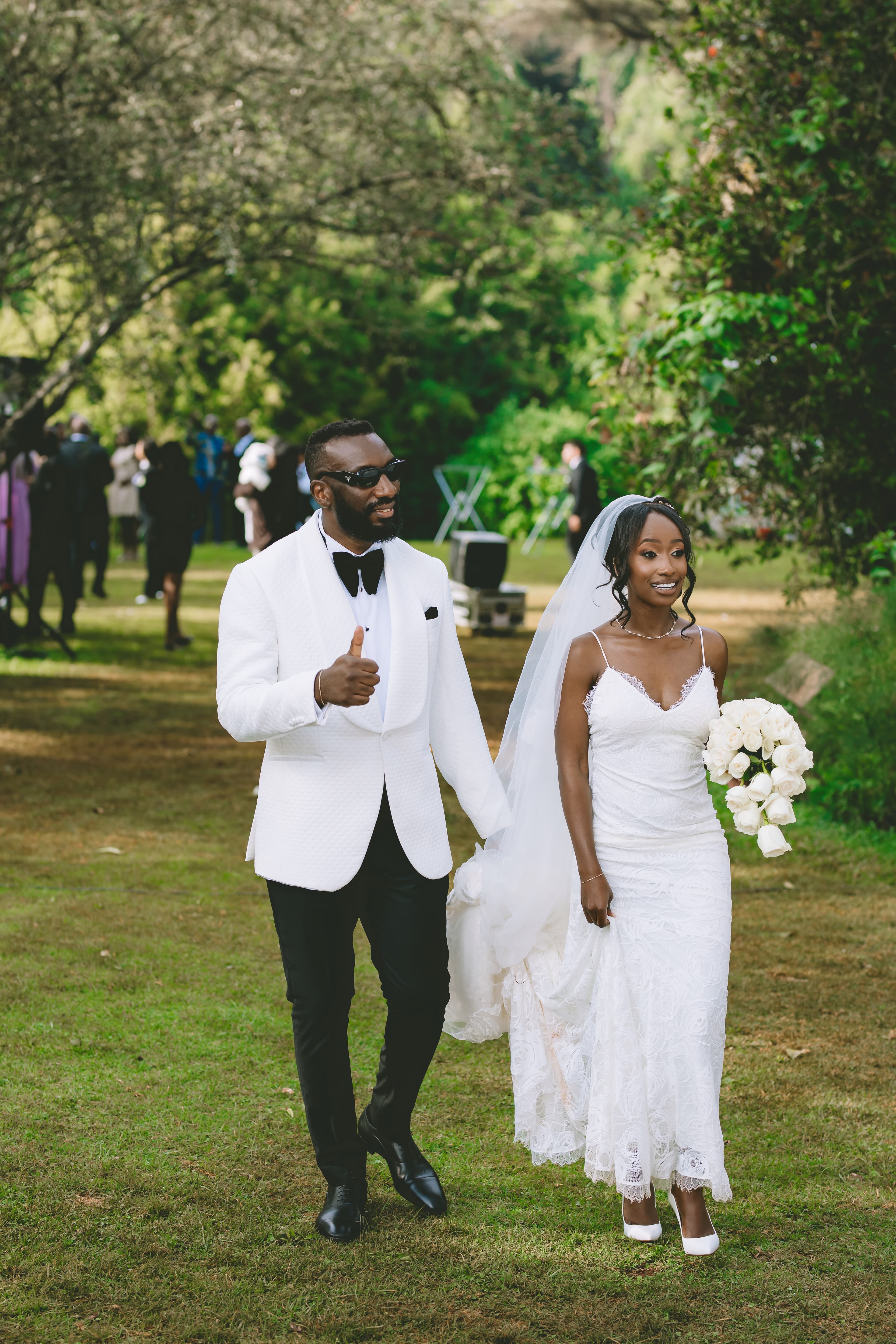 Smiling bride and groom walk outdoors; groom gives a thumbs up, bride holds white roses.