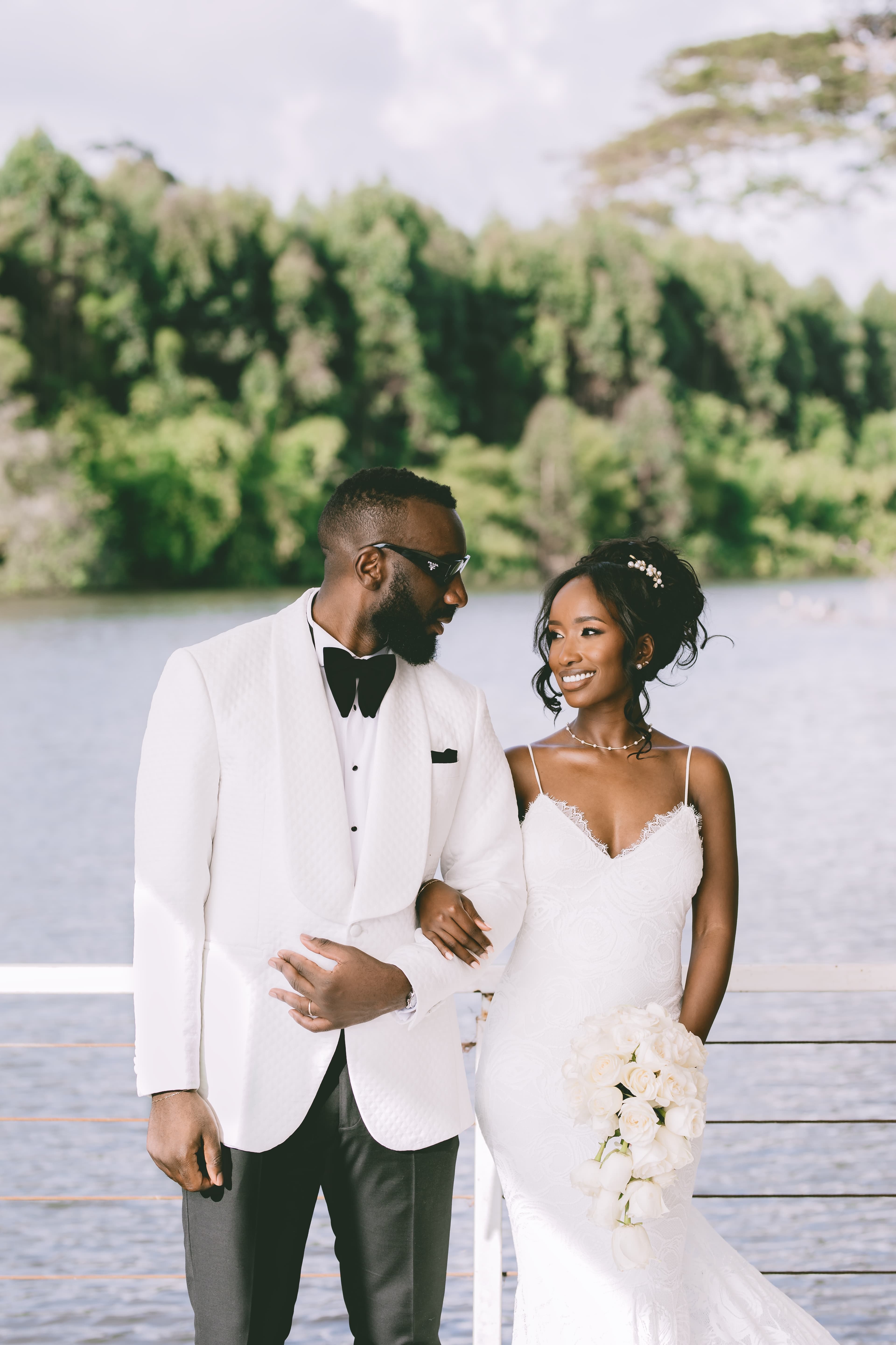 Black bride and groom in white wedding attire look at each other by the water.