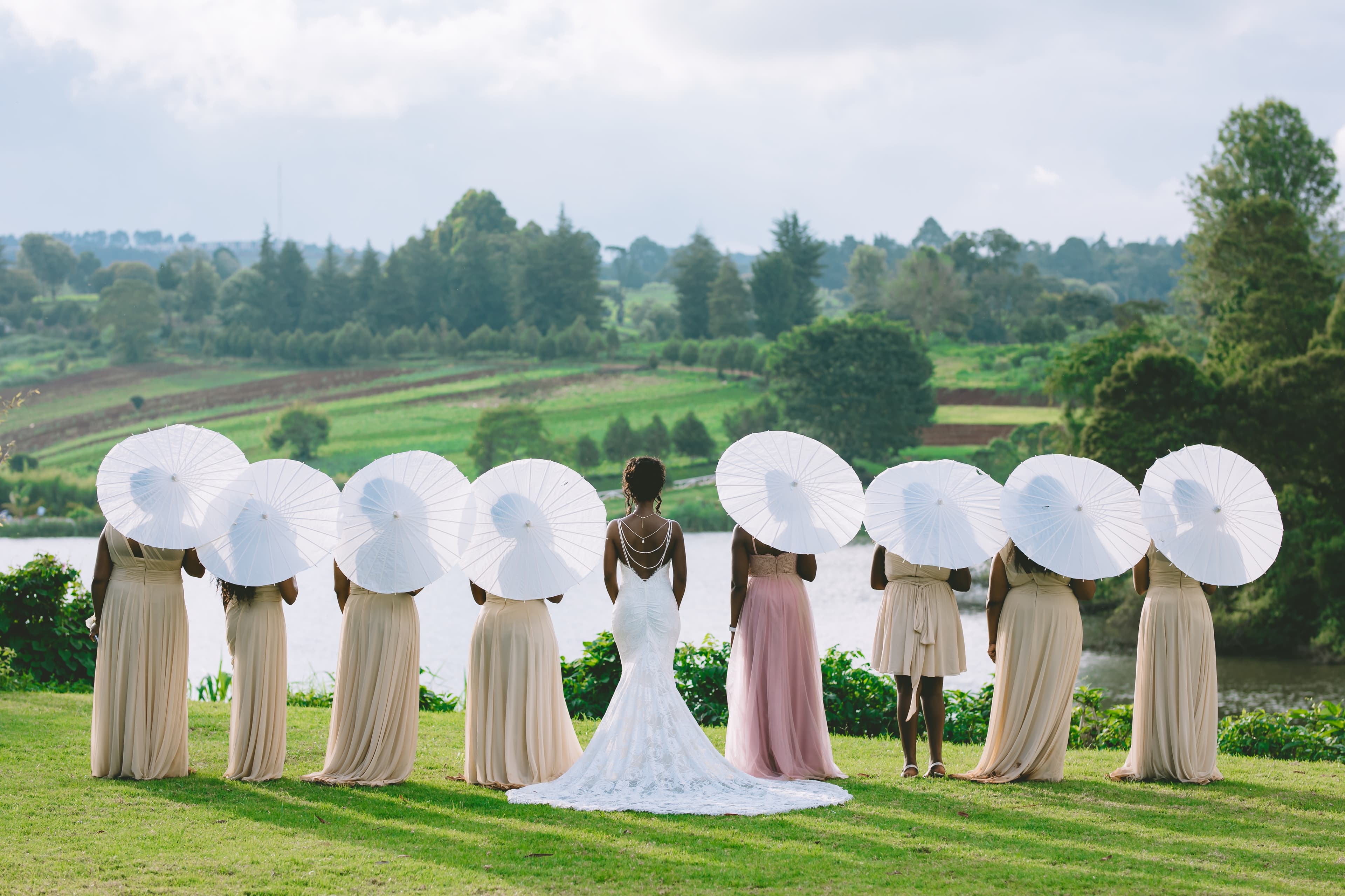 Bride and bridesmaids holding white parasols stand facing a lake and rolling green hills.