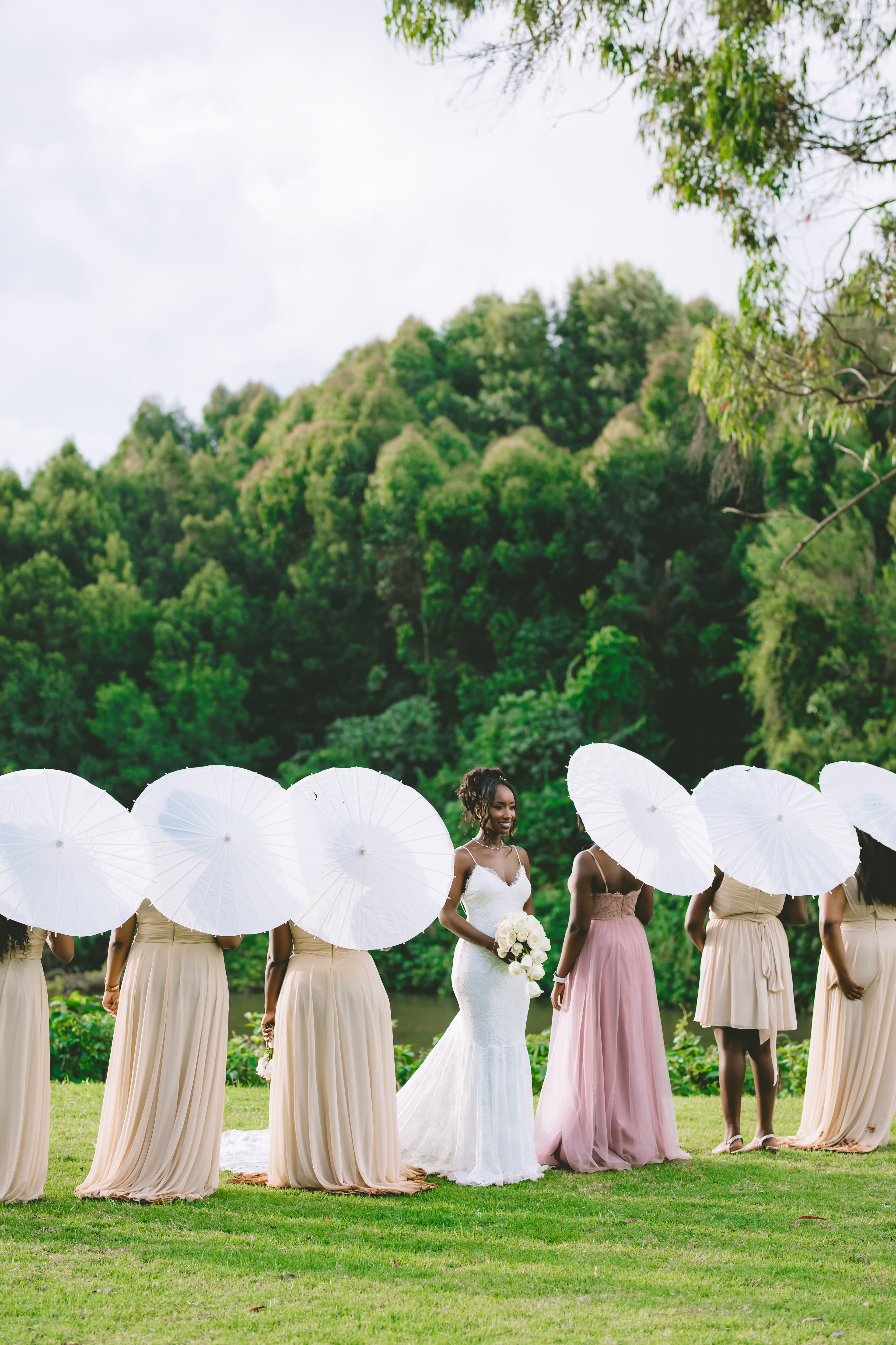 Smiling bride in white lace gown with bridesmaids holding white parasols in a lush landscape.