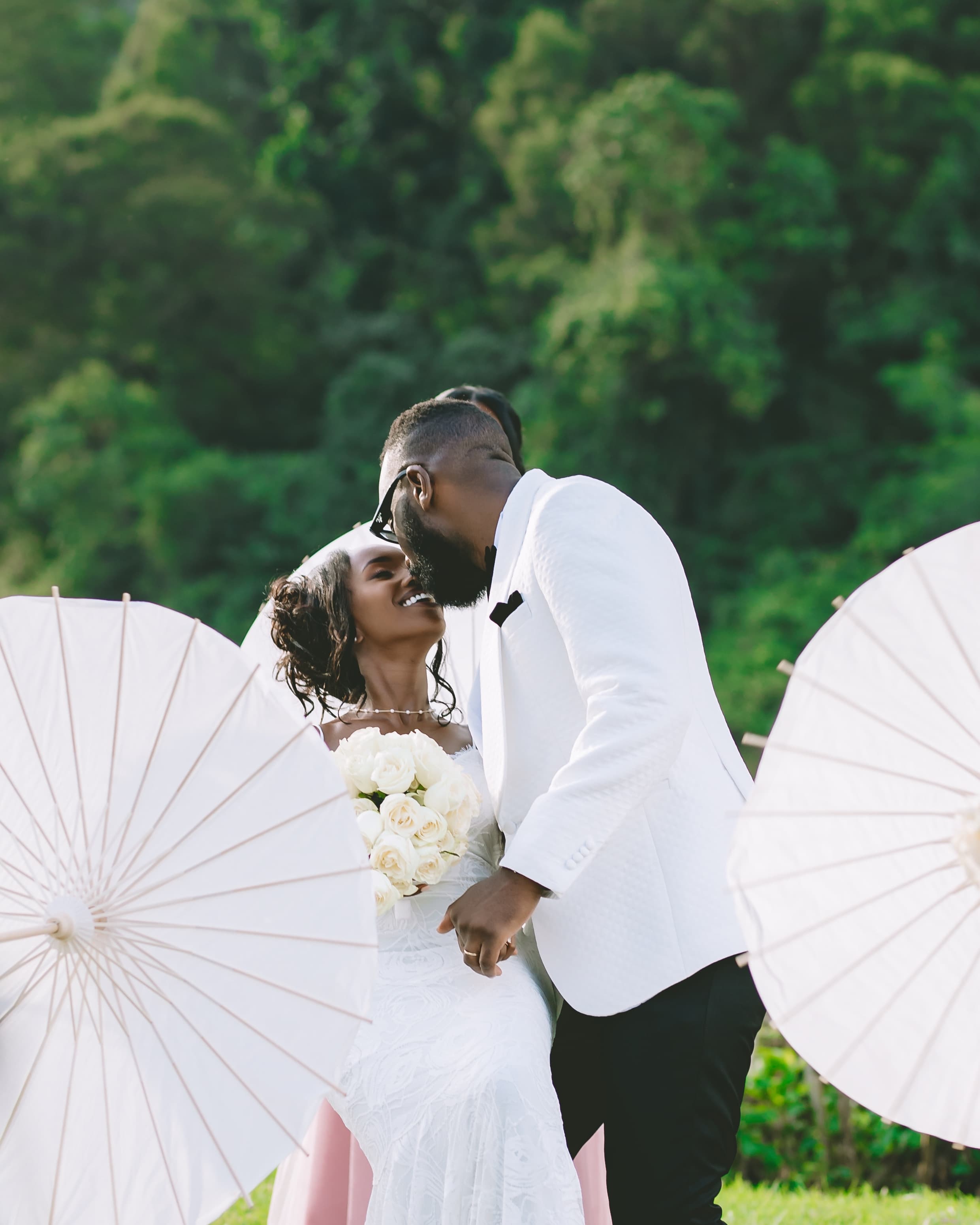 Smiling Black bride and groom lean in for a kiss, framed by white parasols.