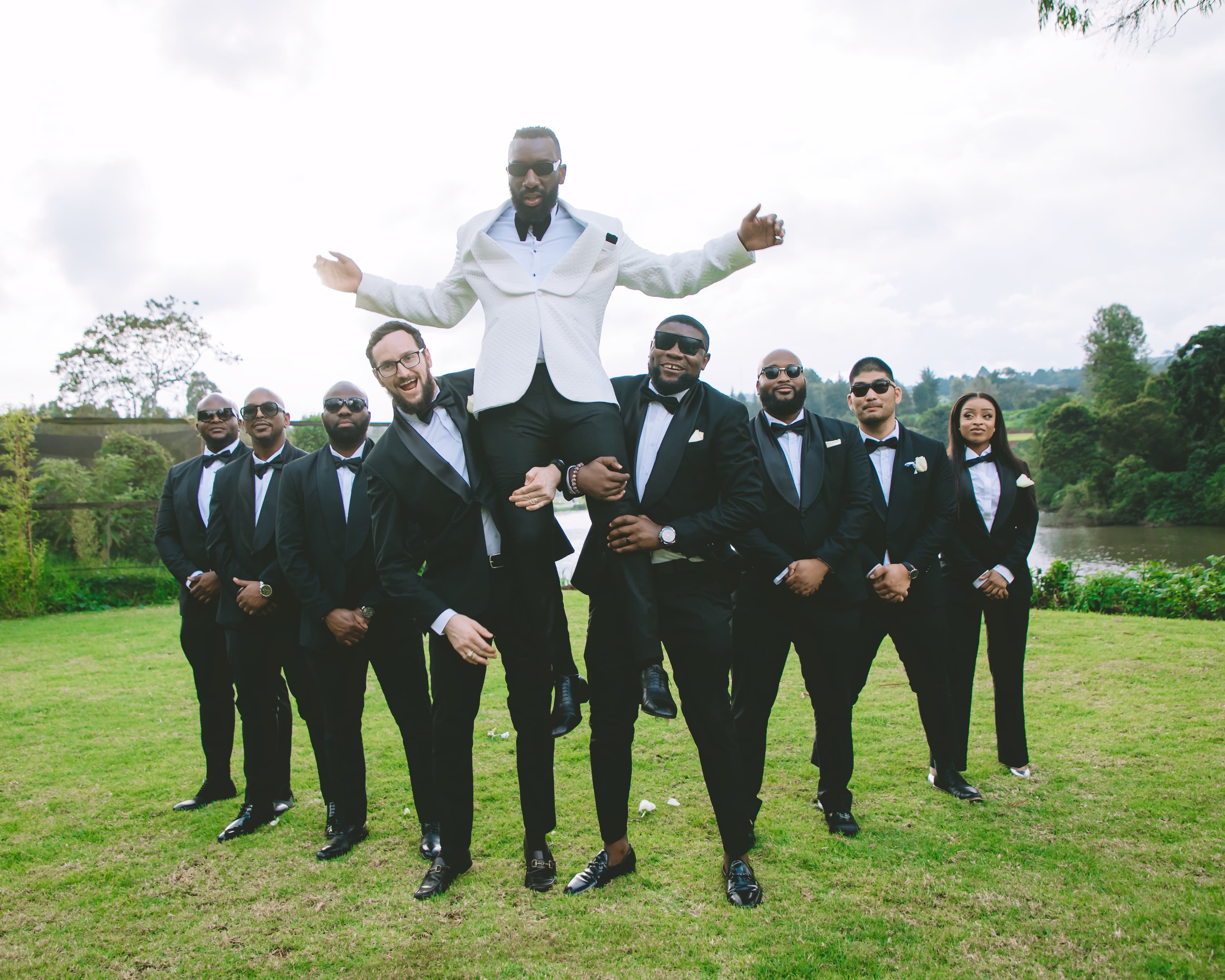Groomsmen in black tuxedos and sunglasses lift a groom in a white jacket outdoors.