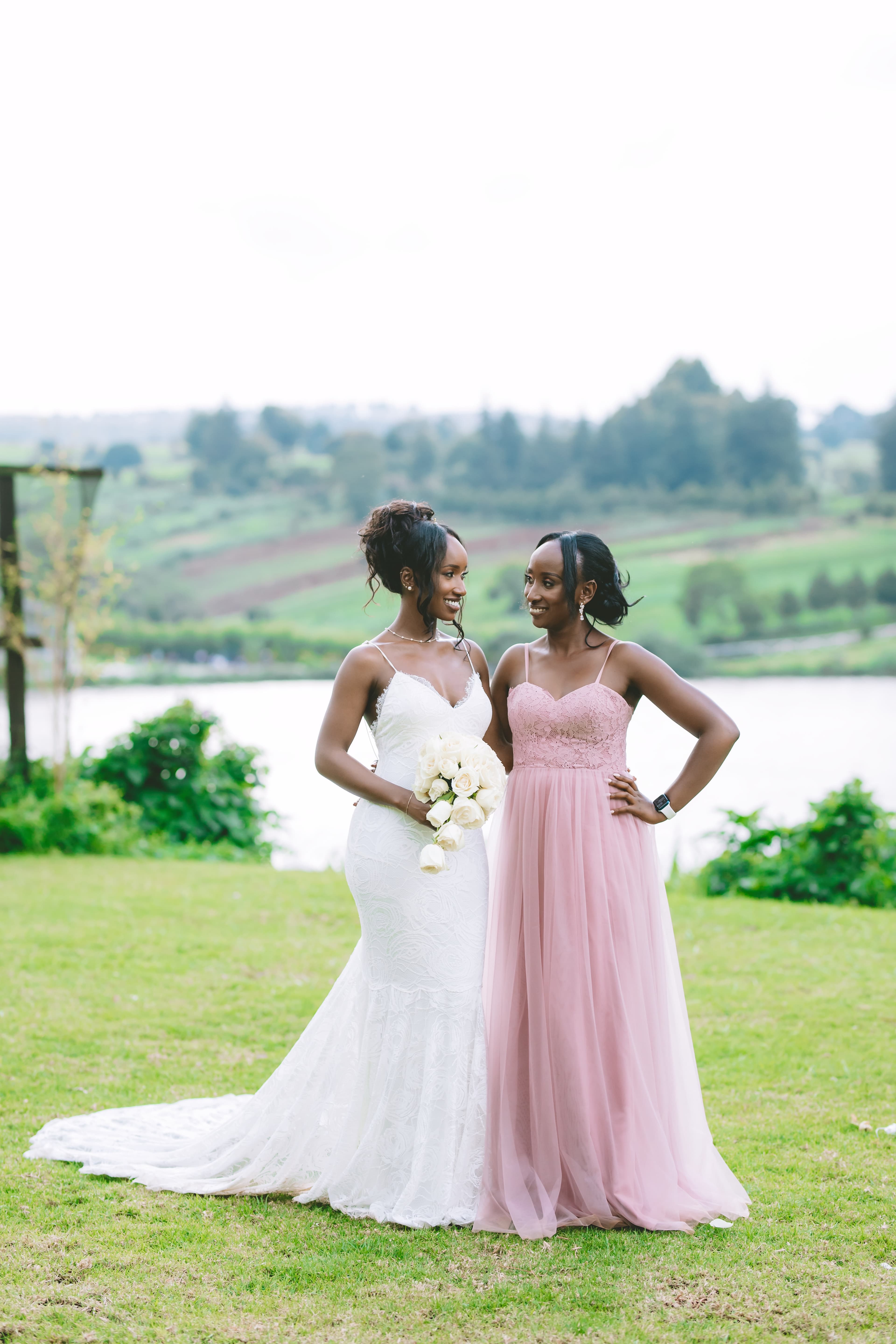 Two Black women, bride and bridesmaid, smiling at each other in a scenic outdoor setting.